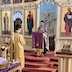 Fr. Aleksey is standing at a podium in from the Royal Doors, addressing  congregants during mission vespers. He is wearing a purple vestment, and from the podium, hangs a long purple cloth with gold fringes around the edges and a gold cross near the bottom. Standing opposite, and facing Fr. Alekey, is an altar boy who is wearing a gold vestment and holding a candle in a tall candle holder.