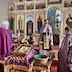 Fr. Aleksey stands in front of the Royal Doors during mission vespers and is facing the congregation. A table in the foreground is covered by a purple cloth that has gold fringes, and there's an icon and two baskets on the table; one is a collection basket, and the other contains booklets and other materials that people can take with them. Two other clergy members are visible in the photo, one one either side of the table. both are looking toward Fr. Aleksey, who appears to be addressing the clergy member who is on the right side of the frame.