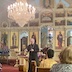 A clergy member, who is wearing a solid, black vestment and large, silver cross on a chain, addresses the congregation during mission vespers. Two congregants are seen seated, in the lower right corner of the frame.