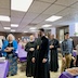 A view of the church hall, following mission vespers. Congregants are standing along the sides of tables that are covered by purple table cloths. Everyone is prayerful and facing towards the lower left. Some white, paper napkins are visible at the corner of the table in the lower left part of the frame, and there's a large, aluminum tin filled with pretzel twists on the corner of the table in the lower right part of the frame.