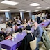 A view of the church hall with parishioners and guests seated at tables and talking. Two tables are visible and run from the lower right corner of the photo to the middle of the right frame. The tables are covered by purple, plastic table cloths.