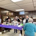 A view of the church hall with parishioners and guests getting food. There's a table with food across the lower left of the frame, and three women are adding food to their plates. Behind them, a group of people wait in lines leading toward two windows, on the left, where kitchen workers are server food. In the lower right corner of the photo, a woman's back faced the camera; she's wearing a teal shirt, and a man, who is standing in front of her but closer to the aforementioned table, is visible over her left shoulder.