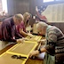 Three of the workers from the previous photo are working at the table, rolling dough into long, snake-like shapes that I'm going to call 'dough ropes'. A man wearing a maroon colored button-down shirt is working next to a woman in a short-sleeved, light red (no, not pink!) shirt on the far side of the table. A woman in a patterned top is working across from them and appears on the right side of the frame, in the lower right corner.