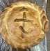 A view of one of the finished paska breads, looking down at the top of the circular loaf. There are braided dough ropes around the circumference of the loaf, and an Orthodox cross embellishes the center. The cross is made out of pieces of the dough ropes. The color of the loaf is golden and slightly more browned on the cross and braids.