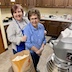 Two woman are standing in the kitchen, posing for the photo. In the foreground, and to their right, is a large, open bag of flour. In front of them, and in the lower right corner, is a silver, industrial-size mixer. In the background, some paska breads that are ready for baking are resting on the counter along with various kitchen utensils and a gallon milk jug that is mostly empty.