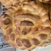 A view of another of the finished paska breads, looking down at the top of the circular loaf. There are braided dough ropes around the circumference of the loaf, and an Orthodox cross embellishes the center. The cross is made out of pieces of the dough ropes. The braids on this loaf are a bit larger than in the earlier photo, and the loaf is darker as well.