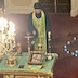 Father Aleksey, looking solemn and with his head bowed, as he blesses the church and parishioners with his silver scensor. He is wearing green vestments and is standing in front of three steps that lead to the Royal Doors behind him. In front of him is a table, covered in a green (but leaning more towards teal) tablecloth, that holds several objects, including a gold cross, an icon, a small, blue prayer book, and some candles. The carpeting on the floor is a deep red, and part of a large chandelier is visible on the left side of the frame. Light blue rectangles, in a somewhat circular pattern, appear on the right side of the frame. They are apparently reflections on the camera lens from the chandelier.