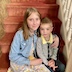 A young girl and a younger boy smile and pose for the camera as they sit on red, carpeted steps that lead up to the choir loft. Somewhat ornate beige posts that are part of a handrail run along the right side of the frame, and a stone-like-patterned, light brown wall runs along the left side of the frame.