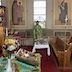 Father Aleksey is standing behind a table that is covered by pussy willows on a white tablecloth. He's wearing green vestments and holding a sliver scensor that he's using to bless the pussy willows. An icon, two white vases, and a red vigil candle holder are on a table visible in the lower left corner of the frame. The table has a green tablecloth, and the icon is rimmed by flowers and pussy willows that match those in the white vases. An altar boy, wearing a gold vestment, is partially visible on the left side of the frame.