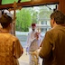 Father Aleksey is standing in the doorway of the church entrance, entering the church. He's facing the camera, and a house and large tree, across the street outside, are visible behind him. He's holding a silver censor in his right hand, and a gold candelabra made up of three candles and a cross. He's wearing white vestmensts and a red cap. Three men stand in front of him. There's a man on the right side holding an icon, and a teenager is on the left side, holding a large gold cross. The third man's ear and a bit of white hair can be seen just past the teen's neck; those are the only parts of the man that are visible.