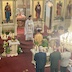 View from the choir loft as Father Aleksey presents the holy gifts. There's an altar boy standing in front of him, holding a candle. There's a table with a white cloth behind the altar boy and then three men standing in a row holding three-pronged candles.