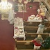 Father Aleksey is holding a prayer book while standing at the corner where two tables with white table cloths meet. The longer of the two stretches out to his right and towards the bottom edge of the photo and runs perpendicular to the pews. The shorter table is in front of the pews and parellel to them. There are two women standing in different row of the pews near the tables, which are covered with Paschal baskets waiting to be blessed. The long table also has a basket of red Easter eggs that will be given to parishioners and guests at dismissal.