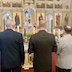 Father Aleksey presents the holy gifts. There's an altar boy standing in front of him, holding a candle. There's a table with a white cloth behind the altar boy and then three men standing in a row holding three-pronged candles. This photo is similar to one described earlier, but the photo was taken from behind the three men, rather than from the choir loft.