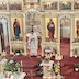 Fr. Aleksey, who is wearing a white vestment, is standing in the archway of the Royal Doors, reading from the Holy Bible as he faces the congregation. The photo is bright and filled with Easter flowers on the three visible icon tables and in various places along the floor, next to walls and/or table, so not really tripping hazards, but lots of pretty flowers.