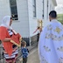 Fr. Aleksey, Matushka Natallia, and a young boy standing outside at one of the corners of the church. Fr. Aleksey is on the right-hand side of the frame; his back is facing the camera, but a sconce of three candles and a gold cross is visible in his left hand. Matushka Natallia is in profile on frame right, and she's holding a red bible with gold lettering. Her hands are draped in a gold-fringed, orange cloth that is between her hands and the bible. She's wearing an orange jacket that almost matches the cloth, and her head is mostly covered by a white cloth. The young boy appears just to Matushka Natallia's left. He's holding an icon and is wearing a patterned shirt in various hues of blue; his back is mostly facing the camera, and his face is not visible.