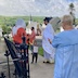 Fr. Aleksey, Matushka Natallia, a young boy and two parishioners standing outside at a different corner of the church. Fr. Aleksey, Matushka Natallia, and the young boy standing are mostly as described earlier, but the boy is now facing the camera. A man in a peach-colored shirt appears in profile and is holding a large, gold cross that is mounted on a pole. A women in a light blue dress appears in the left side of the frame, with her back facing the camera.