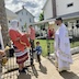 Fr. Aleksey, Matushka Natallia, the man in the peach-colored shirt, and a young boy outside at the same corner of the church as in the previous photo. This photo was taken from a different angle and also shows the back of the rectory and the grassy yard between the church and rectory. In the lower left of the frame, there's a hand holding a gold vessel filled with holy water along with a sprinkling brush for blessing the corners of the church.