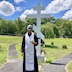 Fr. Aleksey is standing and reading from prayer book while at St. Michael's cemetery. Behind him, and slightly to his left stands a large, silver cross. There's grass, a small tree, and some small plants in the near background, with a circular, concrete path that leads around the cross and gravesite. Further in the distance, at the edge of the cemeteruy, there is a stone wall with larger trees in the background behind the wall.