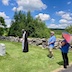 Fr. Aleksey stands before a grave at the middle-left of frame. There's a stone wall about three feet behind him and several trees behind the wall. A woman holding a multi-colored umbrella (for sun!) is looking on from frame right, and there's a man standing a few feet away from her and about a quarter of the way from the right edge of the frame. Both are wearing blue shirts! The sky is bright blue with scattered cumulus clouds.