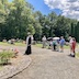 Fr. Aleksey stands just left of center, in the middle of the frame, and he's facing Fr. Dedick's cross, which is out of frame and to the left. Several parishoners stand about eight feet behind him and are hold papers and following along with the service. A few are holding umbrellas. Several graves are visible in the background, with large trees in the very back.