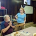 Photo of two people talking in the church hall. The main on the left has white hair and a white beard, and he's seated at a table and is holding his hands together in front of him as though praying, but he's looking at a woman to his left and in the center of the photo. The wom has short, blond hair, and a light blue shirt, and she's standing at the table next to him and talking. There's a plate of food, a church bulletin, and a prayer book on the table in front of the man.