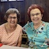 Photo of two seated woman smiling and posing for the camera. The woman on the left has brown hair and an orange and white print shirt, and the woman on the right has red hair and a teal-colored shirt with flowers. Both woman are wearning glasses.