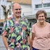 Photo a man and the woman from the previous picture standing outside of the church. The man is on the left side and is wearing a light-black Hawaiian shirt with large flowers, leaves, and birds. Houses are visible in the background.