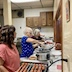 Photo the kitchen in the church hall. Three woman are standing to the left of a counter that runs from bottom center of the photo. They're serving food through windows that are visible on the right side of the picture. Brown cabinets are visible in the back wall, and the counter shows hot dogs on a grill in the foreground and several crock pots. The woman in the lower left corner has brown hair and a orangey-pink shirt. The two women to her left, at the center of the photo, both have gray hair. The one in the middle is wearing a blue shirt with white polka dots, and the woman at the far end is wearing a maroon shirt.