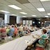 Photo the church hall. Part of two tables are visible, and people are seated at both tables while eating and talking. The table on the right runs from the bottom-left corner to the middle of the right side of the frame.