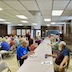 Photo the church hall. A table runs from the bottom center to roughly the center of the photo, and people are seated at the table while eating and talking. Part of a second table is visible to the left of frame.