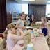 Photo three girls and a boy seated at a craft table that runs from the bottom center to roughly the center of the photo. Two girls are on the left side, and the girl towards the back is looking at the camera. A boy and girl are across the table, at frame right, with the boy closer to the camera. Both are looking at their work.