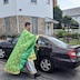 Photo of Fr. Aleksey blessing a different black car. This one is parked on the side street, and the back of the church is visible in the background. The church is on a hill, and the bottom level, where the hall is located, is made up of light-colored stone, while the church agove has white siding. Several windows on both levels are visible along with on the lower level.