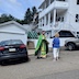 Photo of Fr. Aleksey taking a moment between car blessings to bless a blond woman who is wearing a blue top and white pants. They are outside, standing in an empty parking space between two cars, and white neighbor houses and a white garage are visible in the background.