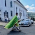 Photo of Fr. Aleksey walking in the street at the intersection near the front corner of the church. The church takes up most of the space to his upper left, and there's a light-blue car in front of him, and towards the right side of the frame. As Fr. Aleksey walks towards the right, his green vestment, with gold embellishments, flies up a bit to the left, almost like a superhero cape.