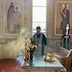 Photo of Fr. Aleksey blessing holy bread that sits on a round, gold platter. The platter has two elevated pedestals on which each of two small, round loaves sit, and the platter also has two gold candleholders and a cross. The left side of the table runs across the bottom frame, and Fr. Aleksey is standing on the opposite side as he blesses the bread with his censer. The table has a light blue cloth with gold trim, and there's a window behind Fr. Aleksey, and two murals on either side of the window. The back side of the table, which appears on the left edge in the center of the frame, has two tall candles, a large votive candle inside red glass with gold embellishments, a gold cross in the center that is only partially visible, and a vase of flowers. Incense smoke rising from the censer appears around Fr. Aleksey with light from the window streaming through the smoke on the left side of the frame.