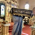 Photo of Fr. Aleksey carrying a large, cloth icon that depicts the falling asleep of Mother Mary, but only the sides are visible. The sides of the icons are deep purple with gold and white flowers in the center, gold lettering near the top, and gold trim along the bottom. Fr. Aleksey is wearing blue vestments, and the altar and iconostasis are visible in the background.