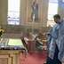 Side view of the aforementioned table, which now appears in the lower left corner of the frame. Fr. Aleksey is standing along the right edge of the frame and is holding a censer and blessing the icon, which has a yellow cloth placed on top, covering the underlying iconography.