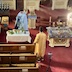 View from the choir loft of Fr. Aleksey standing at a smallish table, covered by a white tablecloth, in the left half of the frame. He's standing on the far side of the table and is blessing several vases of flowers and fragrant herbs that are spread across the table. Two pews, and the top of a third, are visible in the lower left corner of the photo. To Fr. Aleksey's left, in the right half of the frame is the aforementioned table covered by the dormition icon. An alter server stands in the center of the photo, just to the right of the first pew and between the two tables, which are a few feet in front of him.