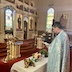 Fr. Aleksey stands in the lower left corner of the frame in front of a table as he reads from a prayer book. The table is covered by a white tablecloth and has several vases containing either flowers or herbs on top along with a gold bowl that's shaped somewhat like a chalice and contains holy water. The background shows the altar and iconostas.