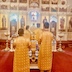 Four clergy members, in two rows, stand behind Fr. Aleksey, who is just inside the Royal Doors at the altar. Their backs are facing the camera as they look towards the altar and iconostas. There is a gold cross and an icon on a table between the two rows of clergy, who are all wearing gold vestments.