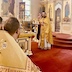 A clergy member is facing the congregation and standing at the base of the steps that lead to the Royal Doors. He's looking slightly to his left at another clergy member who is visible in the lower left corner of the frame. The carpet is red, and a table in front of the clergy member who is standing is covered with a gold cloth that matches the gold vestments worn by the clegy.