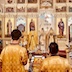 Four clergy members are standing in front of the Royal Doors. Two are holding the Royals and one is holding a cross. The photo is not clearing showing what, if anything, the fourth clergy member is holding. This photo was taken from the center of the church, and two altar servers appear in the foreground with their backs facing the camera. A gold cross and an icon and two votive candles are visible on the table separating the two servers from the clergy.