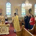 View from the side of the church as Fr. Aleksey standing in front of a table with an icon on it. He is holding a prayer book and praying while two men and a woman stand behind him. The two men are dressed in black, and the one towards the back of the photo is wearing a black vestment and a large sliver cross. The woman on the foreground side is wearing blue pants and a red top that has a white drawing on the front. The drawing is largely out of frame and not easy to make out the details.