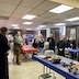 Clergy and parishoners standing the church hall during a prayer before eating. There's a long table, with a blue tablecloth, extending out from the lower right corner, and it's cover with bowls of food. There appears to be bagels (or possibly rolls!), a plate of shrimp, some sort of fruit or vegetable salad, and a few other items including paper plates and condiments. Other tables in the picture have either blue or red tablecloths.