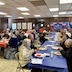 Parishoners and clergy seated at tables in the church hall. Three long tables are visible and are clovered in blue, red, and then another blue tablecloths. The people are eating and talking.