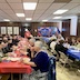 Another view of parishoners and clergy seated at tables in the church hall. Three long tables are visible and are clovered in blue, red, and then another blue tablecloths. The people are eating and talking.