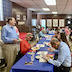 View of one of the blue tables, where parishoners and clergy are eating and talking. A man in a blue shirt and tan pants is standing at the left edge of the frame and is conversing with a woman seated at the table.