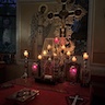 Picture of the altar table lit by candle light. The table is covered by red cloth, and there are three objects on the foreground side of the table. The item on the left is only partially visible but appears to be an icon. There's an ornate bible in the center, and a cross on the right. The back half of the table has several candles on the sides and back of a model church that is enclosed in what appears to be a glass enclosure. There's a large, ornate cross behind and towering above the enclosure. Stained glass windows and a life-sized icon of a person, painted on the wall, are visible in the background, with a bit of blue, evening light showing through the top of one of the windows.