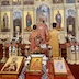 View of the opening archway for the Royal Doors along with the two side doors. Fr. Aleksey's head and right shoulder are visible through the archway as he stands at the altar, with Archbishop Mark standing behind him. There are two altar servers behind and to either side of Archbishop Mark. There's also a table in the foreground displaying three icons, with a cross that's surrounded by small flowers and a red vigil candle on either side of the cross.