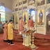 A view of the previous photo but shown from a side angle. Fr. Aleksy can now be seen holding an ornate bible, and the aforementioned table is out of frame. Light is streaming in through a stained glass window on the left frame, as well as through an altar window seen through the archway.